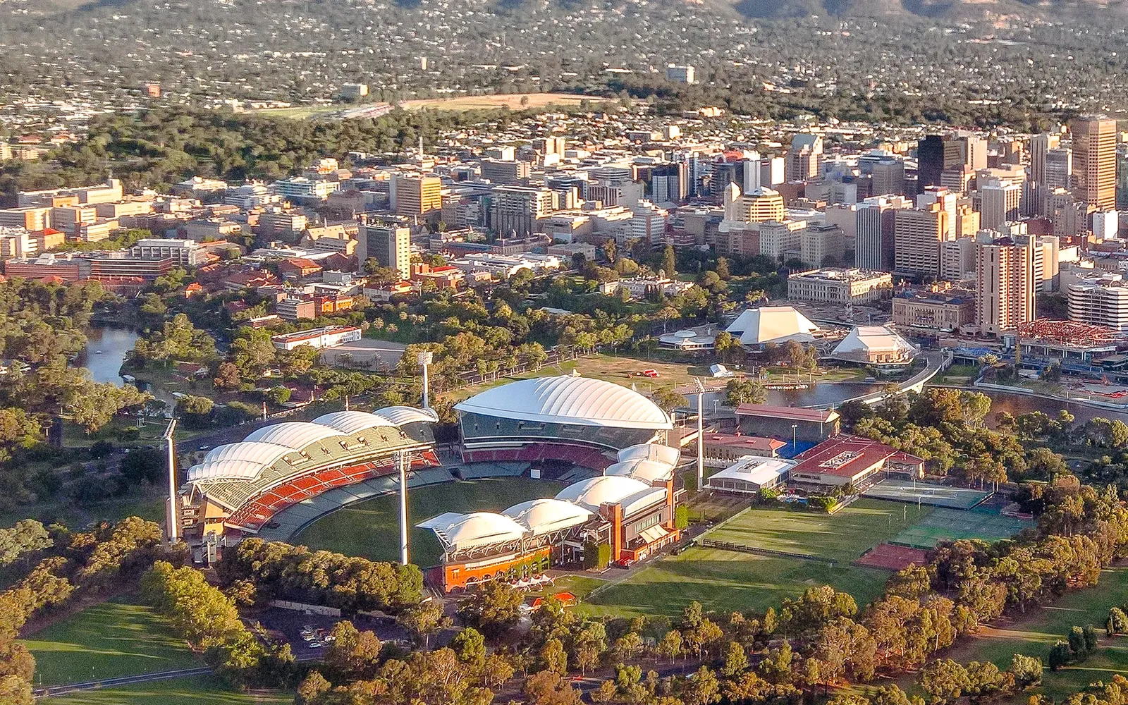 สนาม Adelaide Oval จากมุมสูง สนามคริกเก็ตสีเขียวเข้มล้อมรอบด้วยอัฒจันทร์โมเดิร์นและต้นไม้ใหญ่ร่มครึ้ม มีเส้นขอบฟ้าเมืองเป็นฉากหลัง