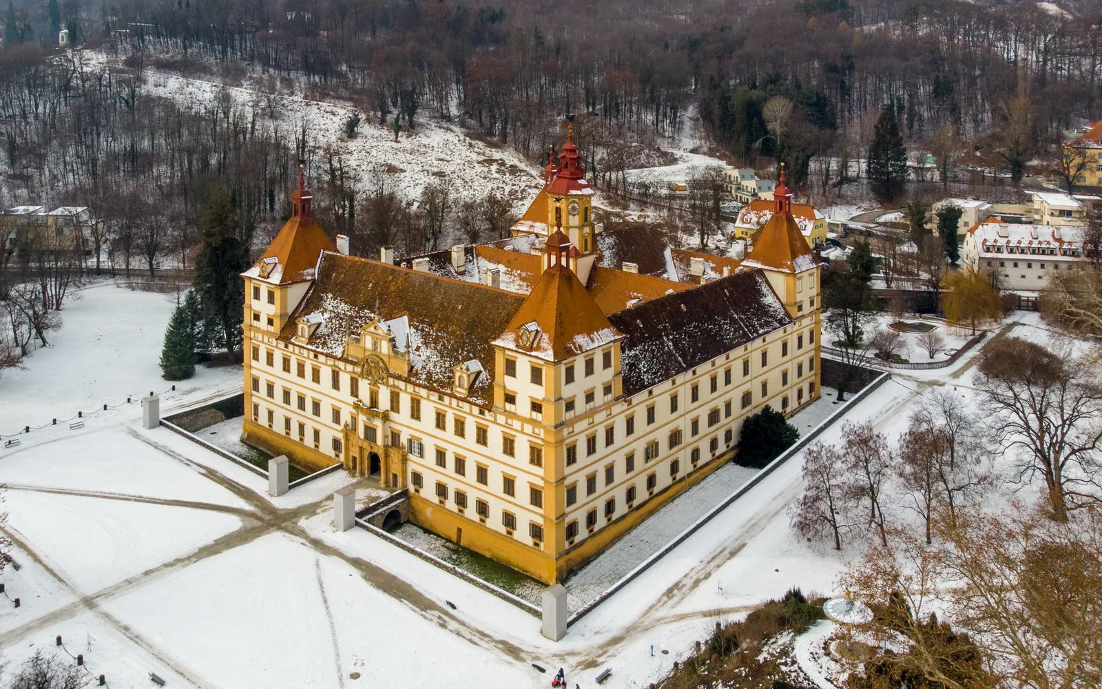 ด้านหน้าวัง Eggenberg ตึกสีเหลืองบาโรกขนาดใหญ่มีหอสูงสี่มุม ต้นไม้สูงเรียงตลอดทางเดินกลาง สนามหญ้าเขียวสดกว้าง