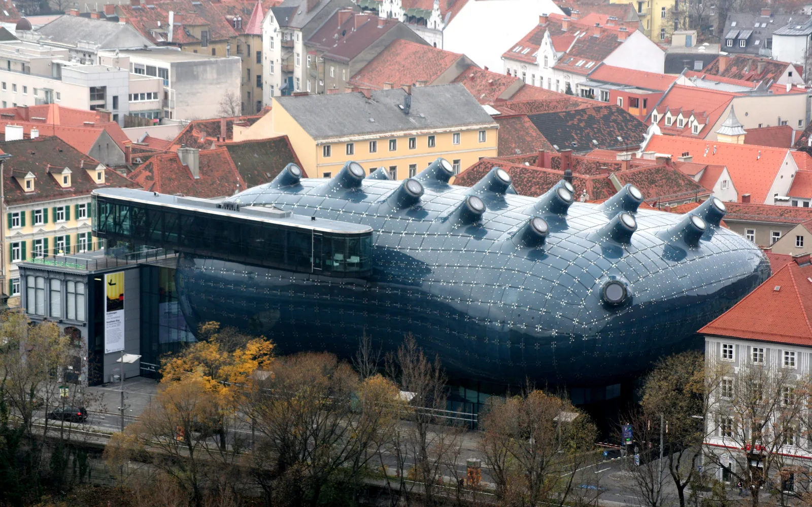 อาคาร Kunsthaus Graz รูปทรงโค้งมนสีน้ำเงินเข้มเหมือนหอยทากยักษ์ตั้งอยู่ท่ามกลางอาคารเก่าสีแดงรอบข้าง