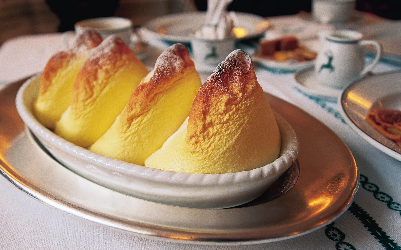 Salzburger Nockerl ในจานขาวลึก ฟูฟองสีเหลืองทองสามยอดเหมือนภูเขาสามลูก โรยน้ำตาลผงสีขาวและราดซอสแอปริคอตสีส้ม