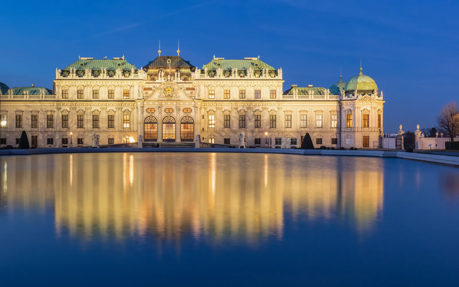 The Upper Belvedere reflected in the rectangular pool out front, its green-and-gold Baroque roof set against a deep blue afternoon sky, parterre flower beds lining both sides