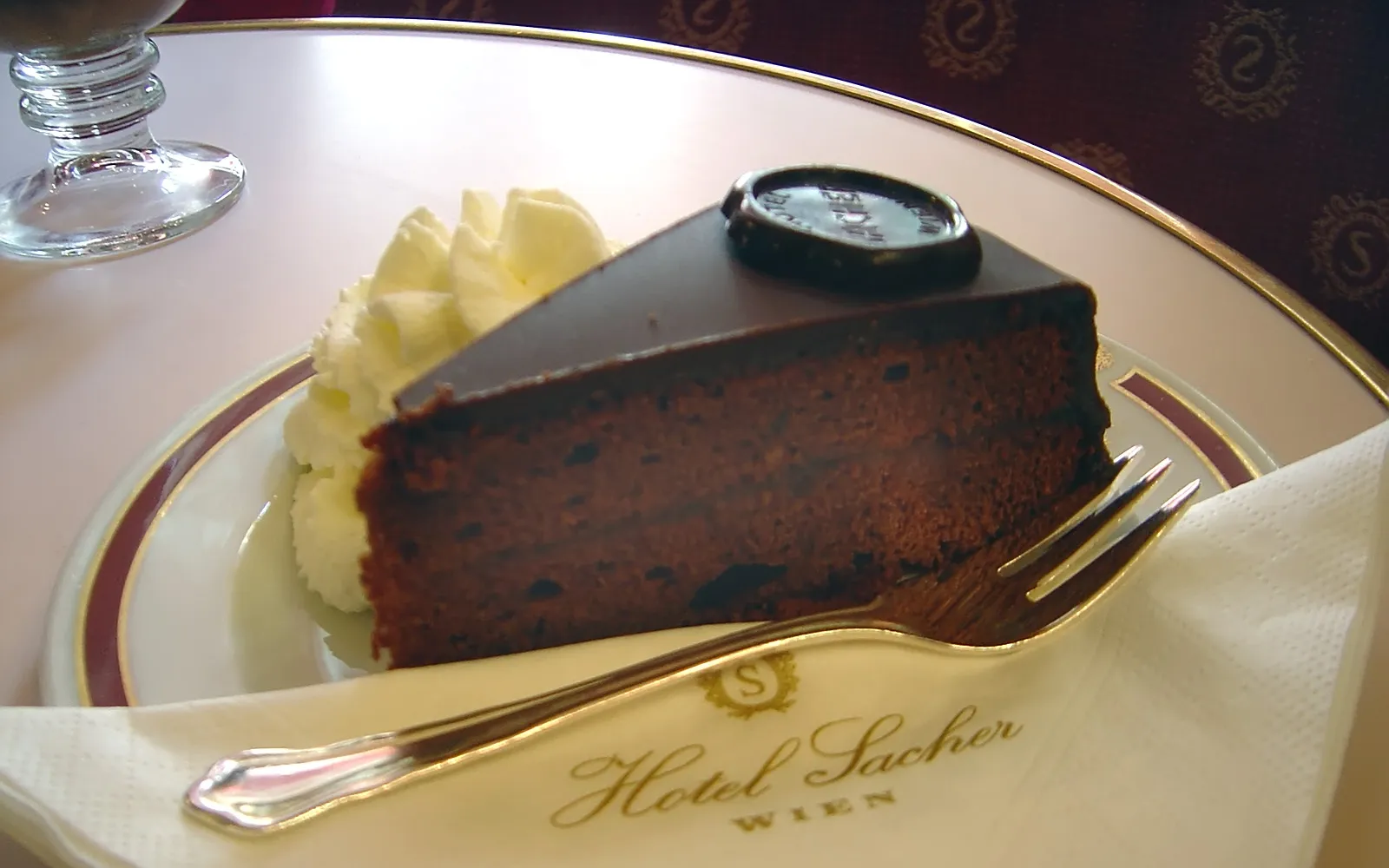 ชิ้น Sachertorte เค้กช็อกโกแลตหนาแน่นเคลือบกานาชช็อกโกแลตมันวาว แยมแอปริคอตระหว่างชั้น เสิร์ฟบนจานขาวพร้อมวิปครีมฟู