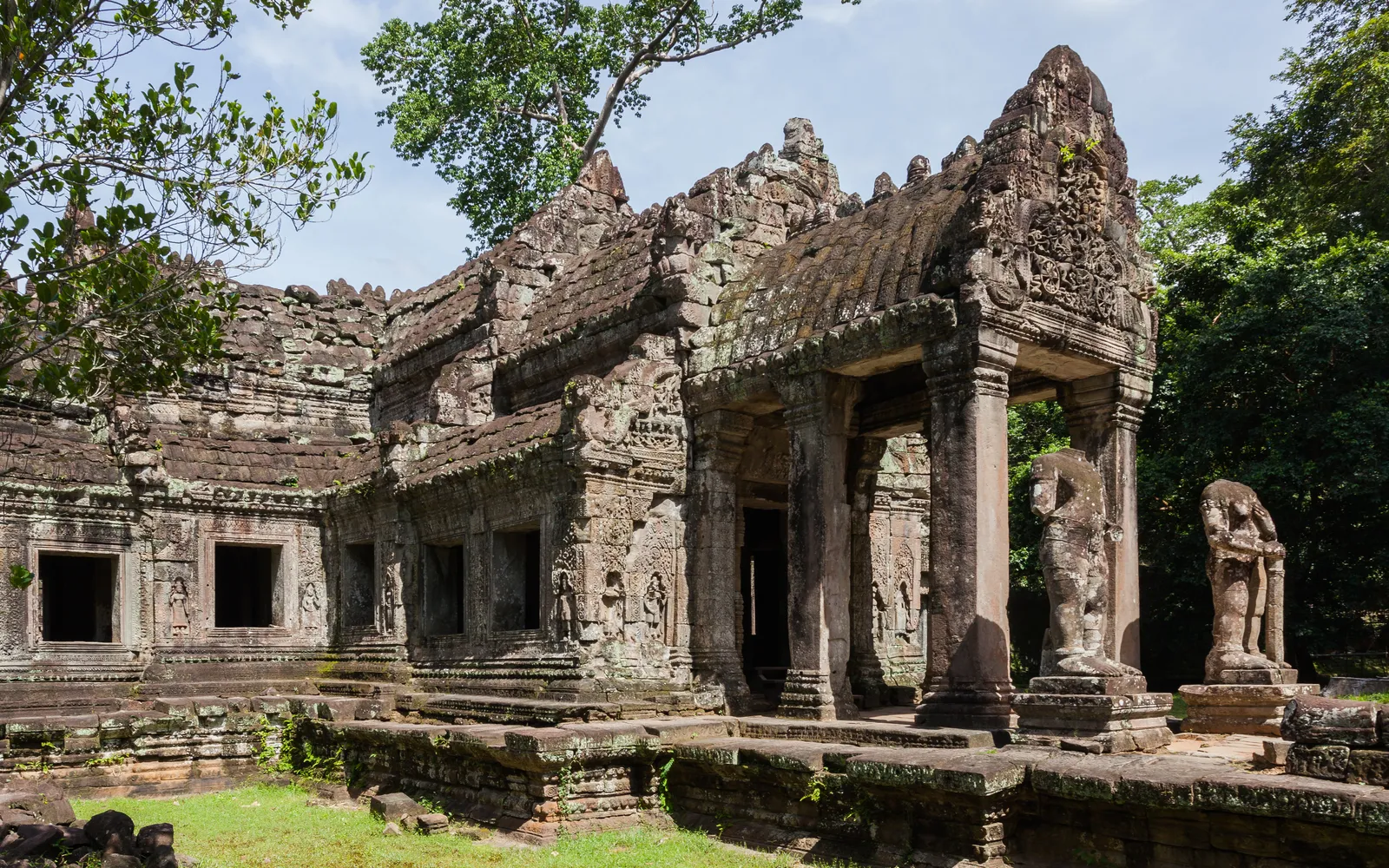 ทางเดินในปราสาทพระขรรค์ เสาหินทรายเรียงเป็นแนวยาวสองข้างทาง รากไม้ใหญ่พาดผ่านประตูโค้ง แสงกรองผ่านรูรั่วบนหลังคา