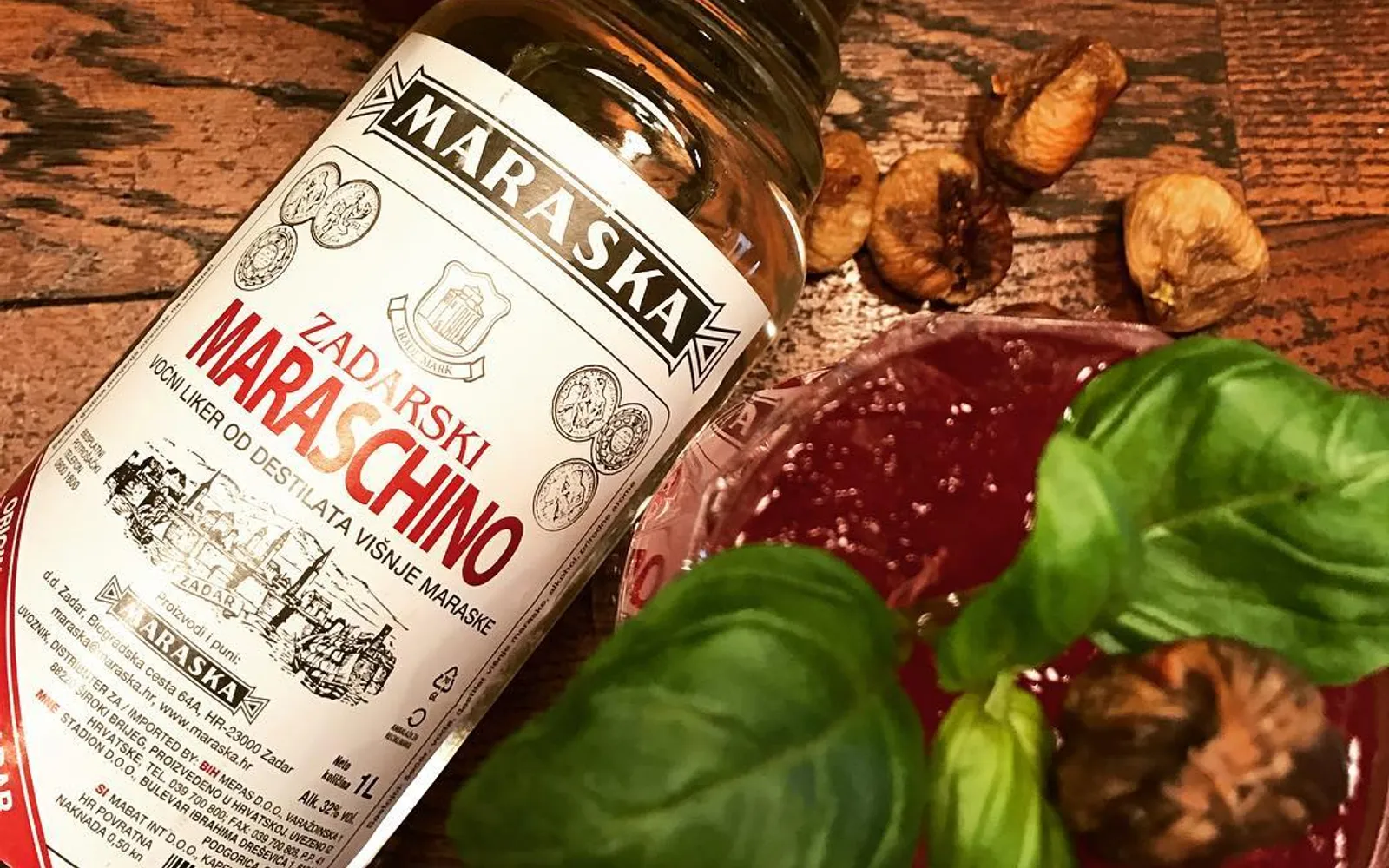 ขวดเหล้า Maraschino ทรงสูงบรรจุในกล่องหวายขาว วางบนโต๊ะหินพร้อมแก้วเหล้าขาวใส