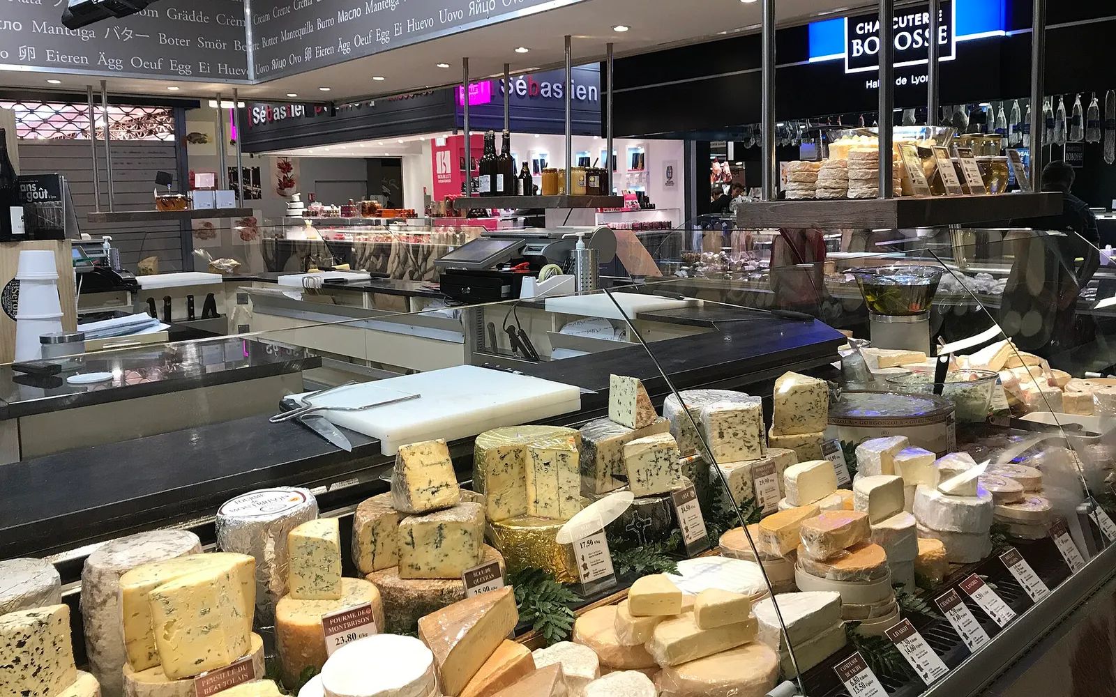 ภายในตลาด Les Halles Paul Bocuse แถวร้านค้าเนื้อ ชีส ซีฟู้ดและขนมเรียงสองข้างทางเดิน แสงสีขาวสดใส ลูกค้าเดินจับจ่ายหน้าตู้กระจกโชว์สินค้าสด