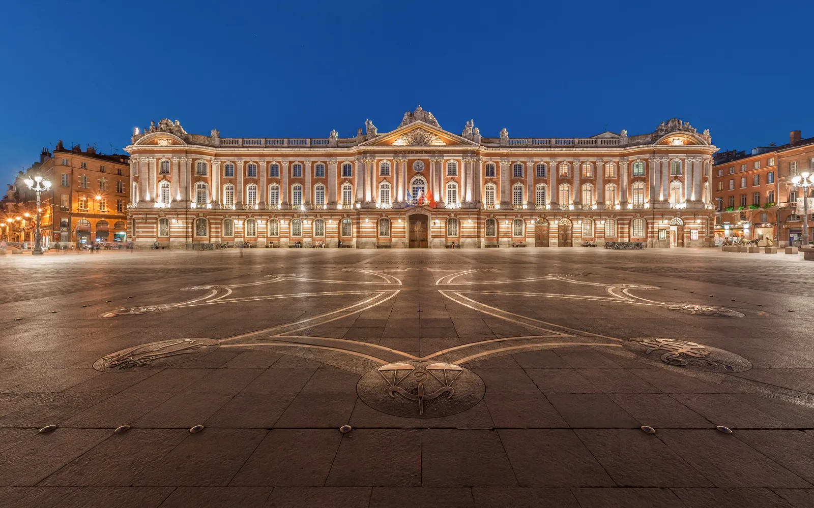 จัตุรัส Place du Capitole ตูลูส อาคารศาลาว่าการอิฐสีชมพูสง่างามตั้งตระหง่าน ผู้คนนั่งจิบกาแฟใต้ร่มเงาริมจัตุรัสกว้าง