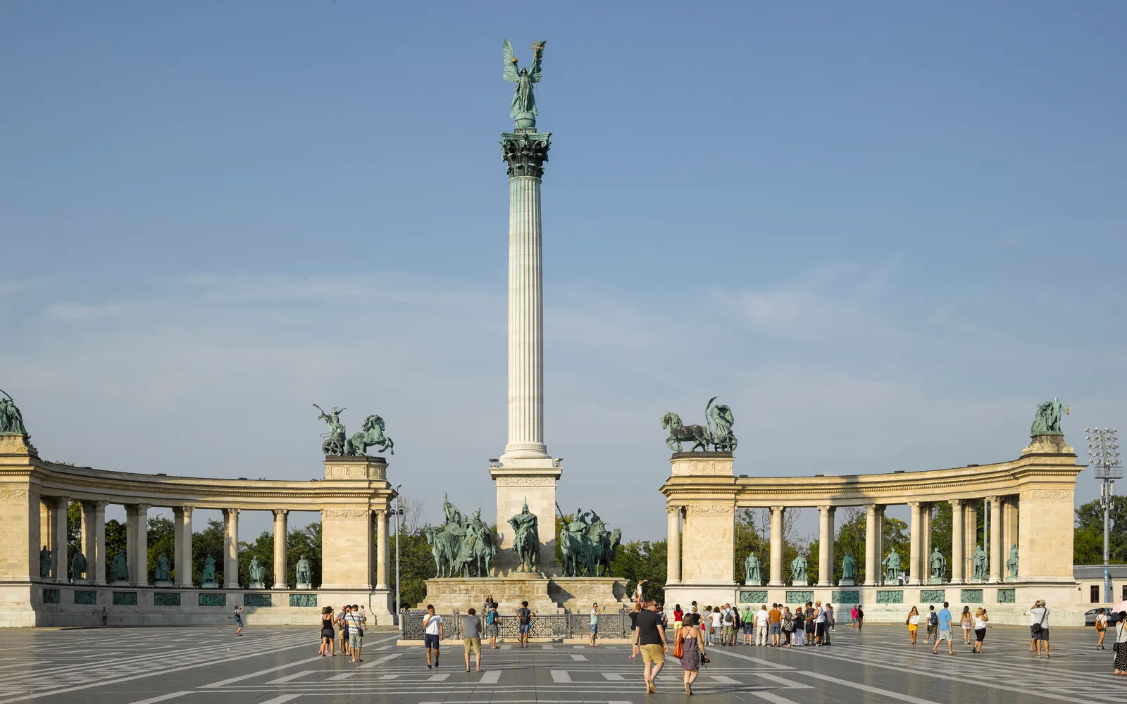 จัตุรัส Heroes Square บูดาเปสต์ เสาหินสูงตรงกลางมีเทวดาถือตราสัญลักษณ์ประเทศ ซุ้มรูปปั้นกษัตริย์และวีรบุรุษฮังการีทั้งสองข้าง ท้องฟ้าสีฟ้าใสเป็นฉากหลัง
