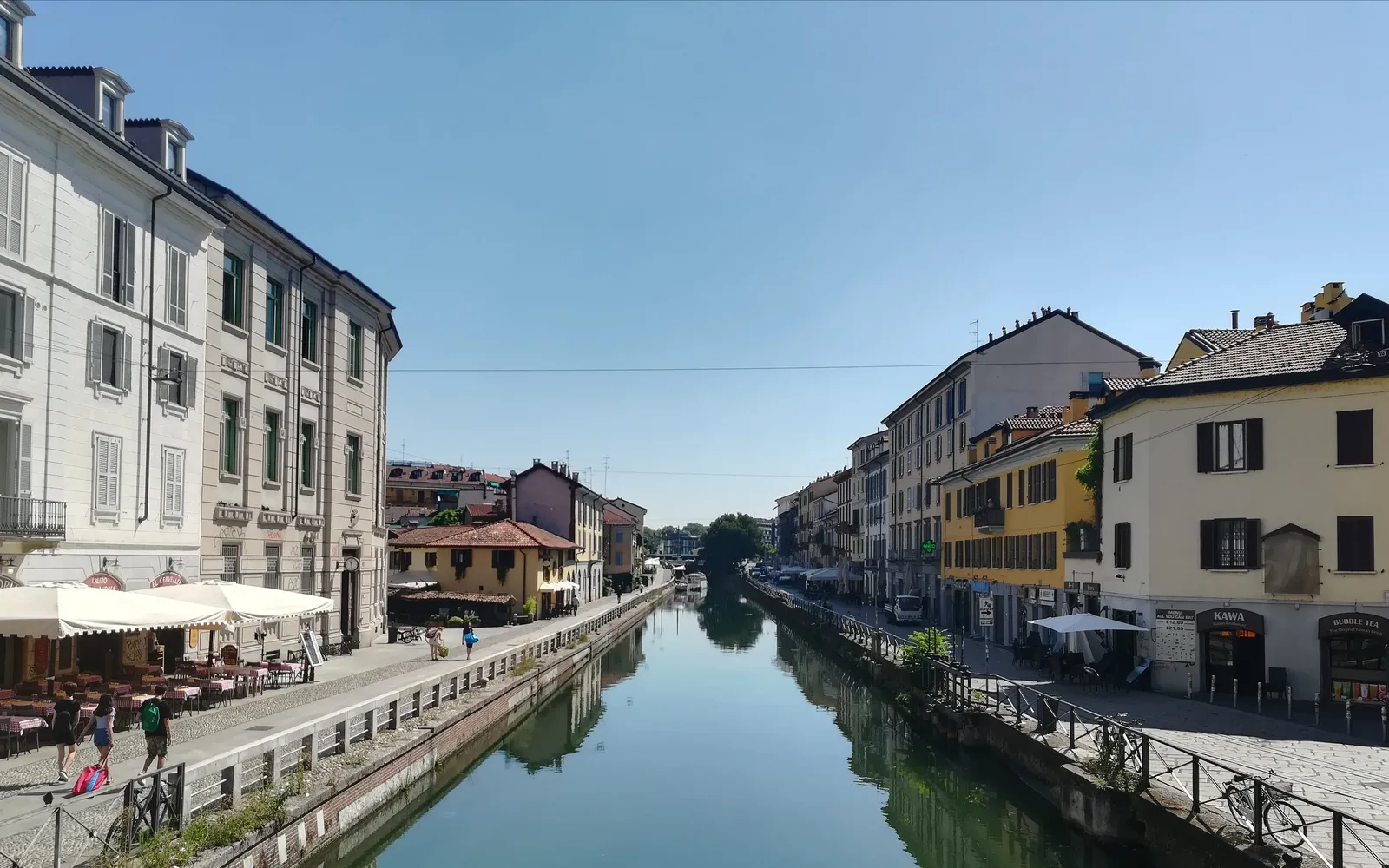 ริมคลอง Naviglio Grande ในย่านนาวีลีมิลานยามค่ำ แสงไฟจากบาร์และร้านอาหารสะท้อนบนผิวน้ำ นักท่องเที่ยวและชาวมิลานนั่งเล่นริมน้ำ