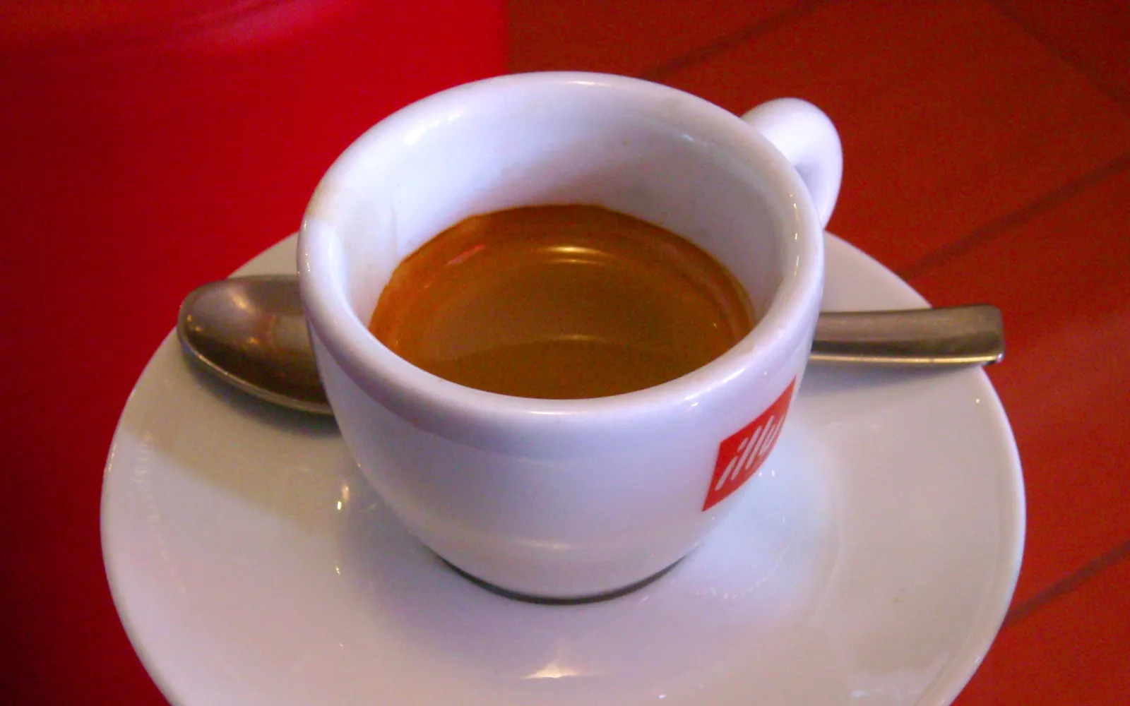 กาแฟเอสเปรสโซเนเปิลส์ในถ้วยพอร์ซเลนขาวเล็ก สีน้ำตาลเข้มเหลือบทองครีมา ควันบางๆ ลอยขึ้น วางบนจานรองพร้อมน้ำตาลก้อนหนึ่ง