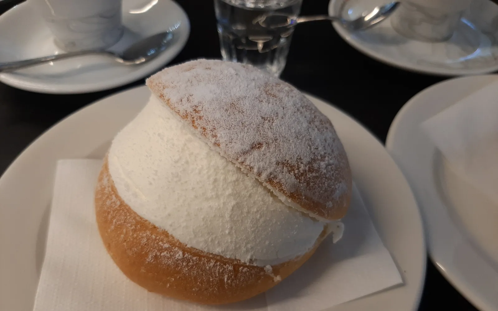 A maritozzo on a white paper plate — a soft golden-brown bun split in half and packed with fresh whipped cream rising higher than the bun itself