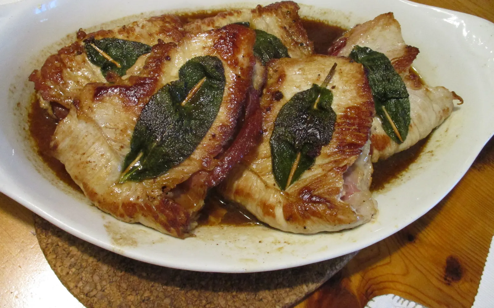 Saltimbocca in a small iron pan on a wooden table — golden-brown veal slices folded with prosciutto and fresh sage leaves, in a glossy white butter sauce