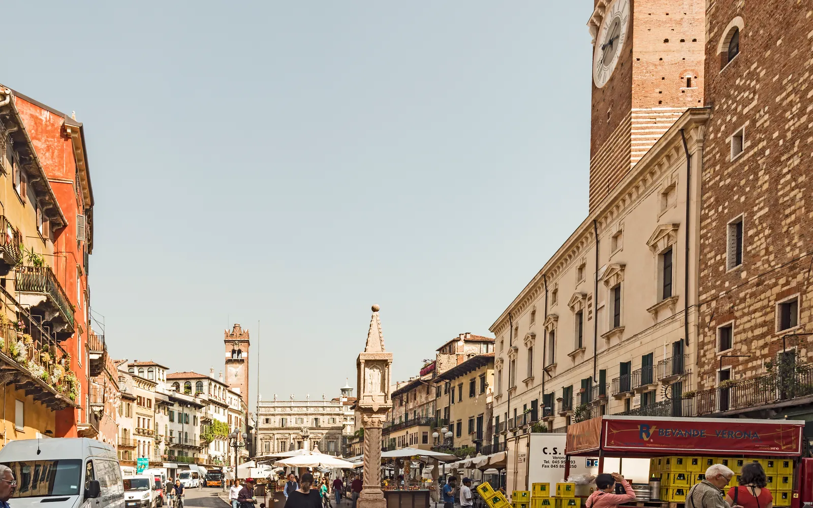 จตุรัส Piazza delle Erbe เวโรนายามสาย ร้านค้าร่มสีสันเปิดขายของกลางแจ้ง อาคารเก่าแก่สีพาสเทลล้อมรอบ หอคอย Torre dei Lamberti ตั้งตระหง่านอยู่พื้นหลัง