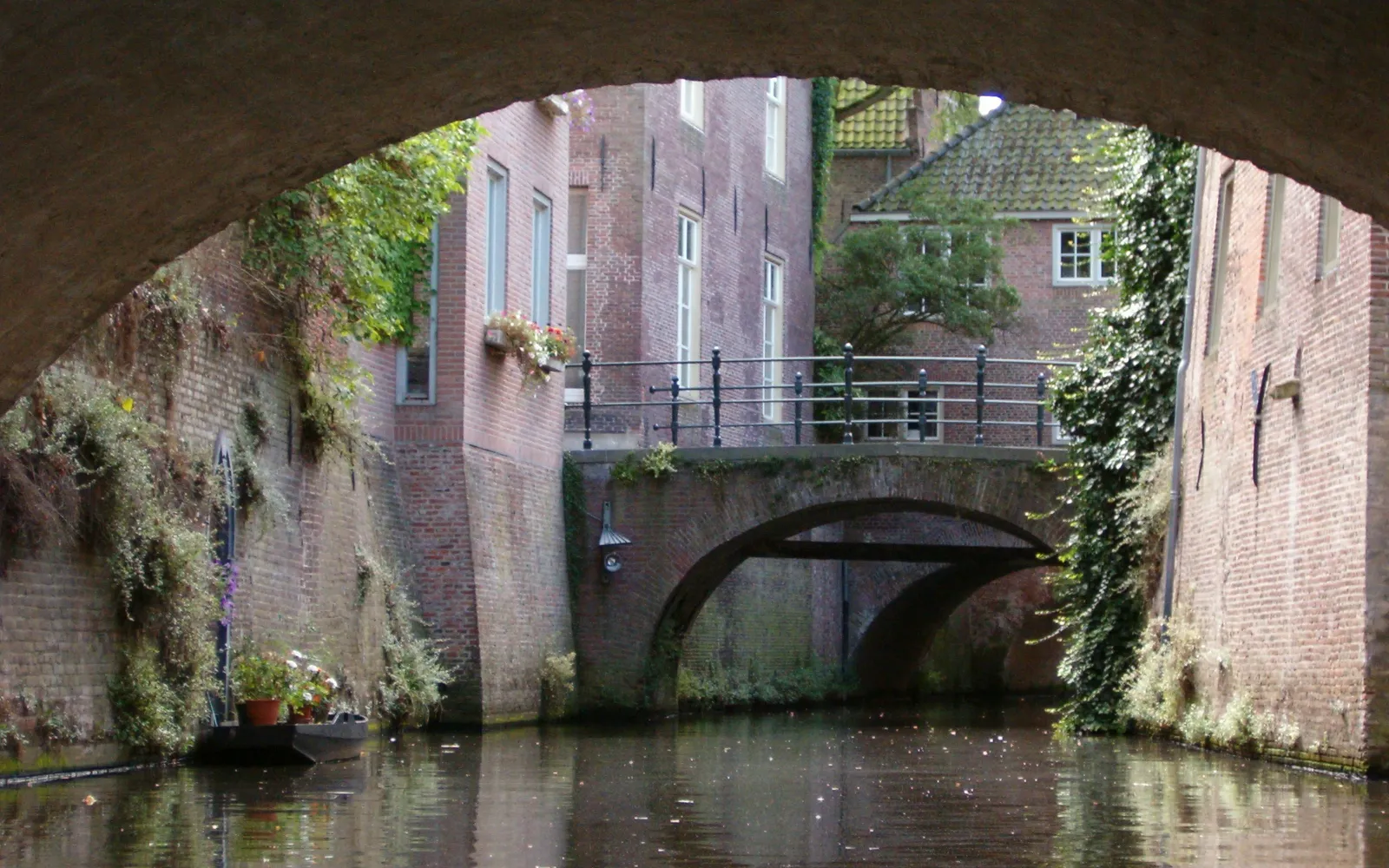 เรือเล็กล่องผ่านอุโมงค์อิฐโบราณใต้อาคารเดน บอช ฝ้าเพดานอิฐแดงโค้งต่ำเพียงไม่กี่เซนติเมตรเหนือหัว น้ำคลองสะท้อนแสงสลัวในอุโมงค์แคบ