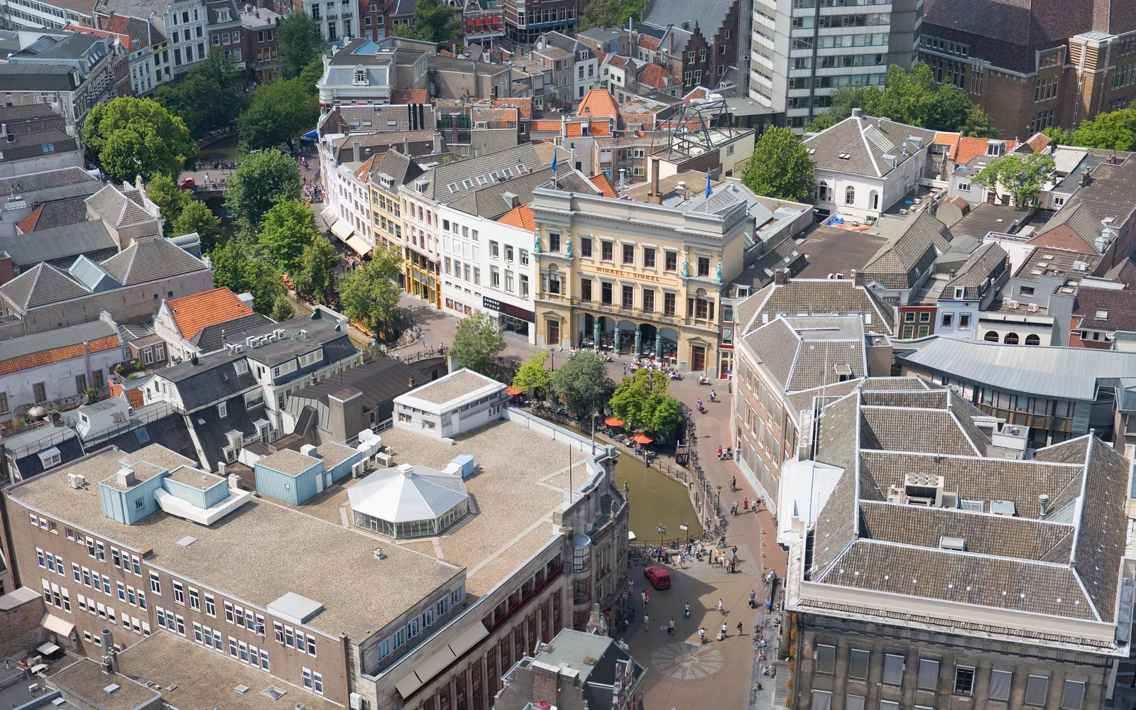 คลอง Oudegracht ยามเย็น ร้านอาหารและบาร์เปิดในช่องอิฐโบราณใต้ระดับถนน เรือจักรยานและเรือยางลอยอยู่บนผิวน้ำสีเขียว