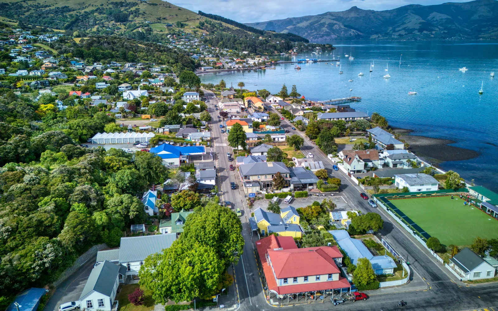 ท่าเรือ Akaroa บน Banks Peninsula ไครสต์เชิร์ช อ่าวน้ำสีน้ำเงินล้อมด้วยเนินเขาสีเขียว บ้านไม้สไตล์ฝรั่งเศสเรียงริมถนนเล็ก