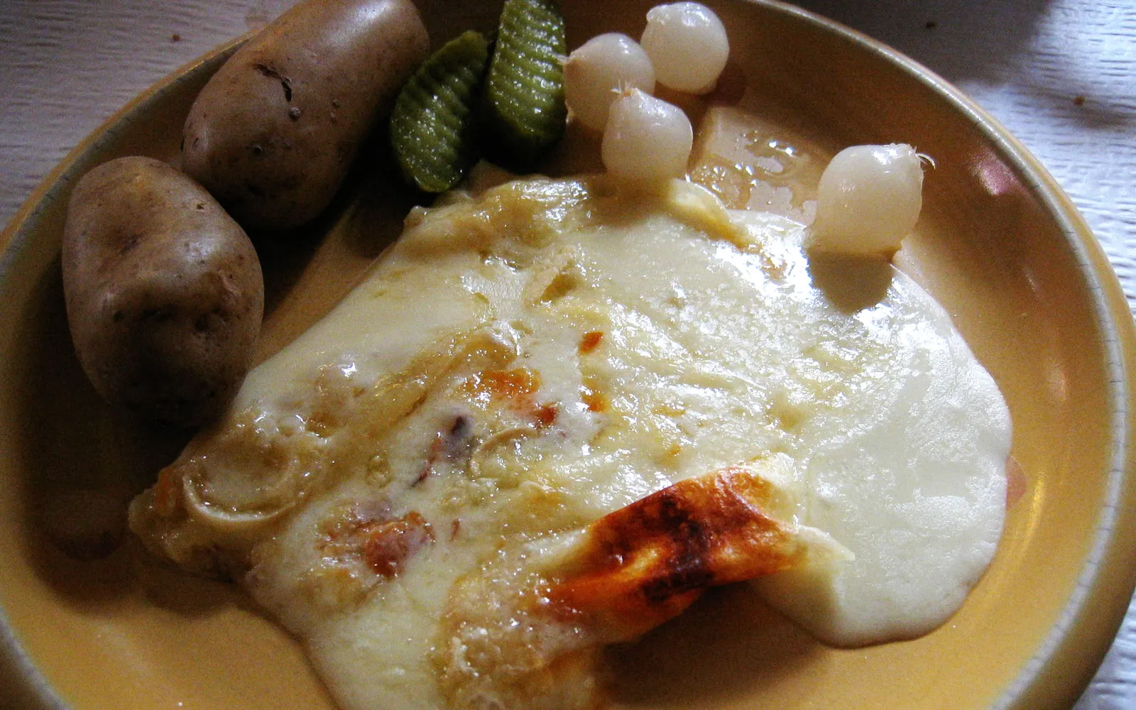 ชีส Raclette ครึ่งวงกลมอยู่ใกล้ความร้อน ผิวชีสละลายเป็นฟองสีทองก่อนถูกขูดลงบนมันฝรั่งต้มและหัวหอมดอง
