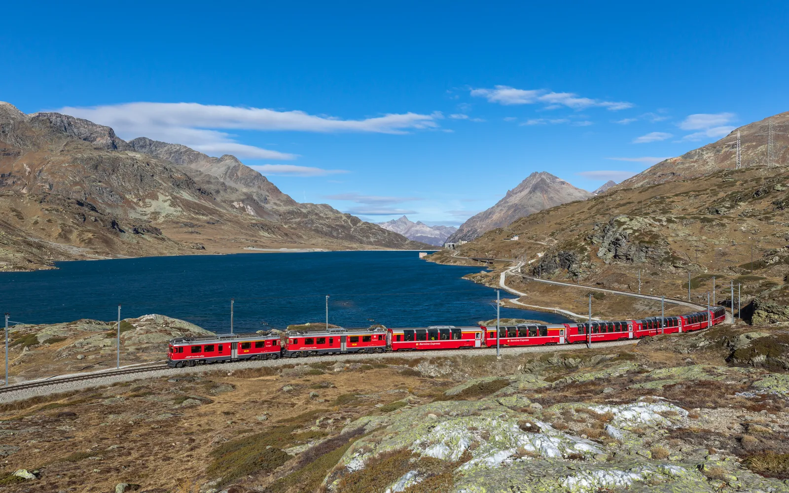 รถไฟ Bernina Express หัวสีแดงวิ่งบนสะพานหินโค้ง Brusio ท่ามกลางทุ่งหญ้าเขียวสดและหมู่บ้านเล็กๆ ในหุบเขาสวิส-อิตาลี