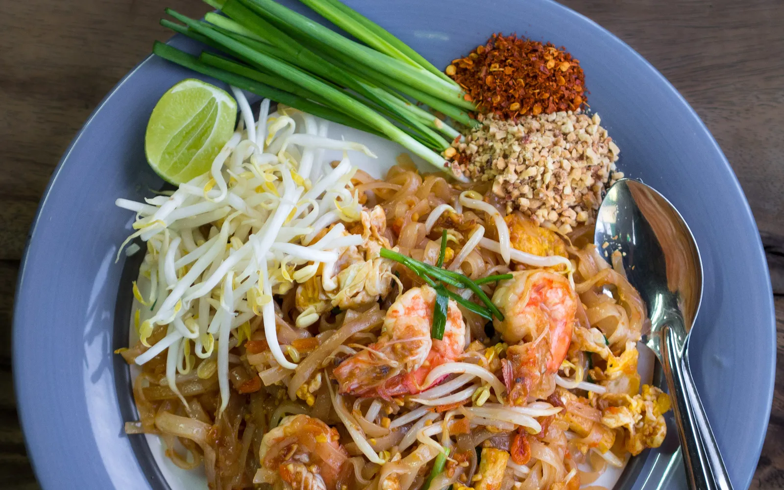 ผัดไทยกุ้งสดในกระทะเหล็กหล่อ เส้นก๋วยเตี๋ยวผัดกับไข่ถั่วงอก ต้นหอม กุ้งสด โรยถั่วลิสงบด มะนาว และพริกป่น