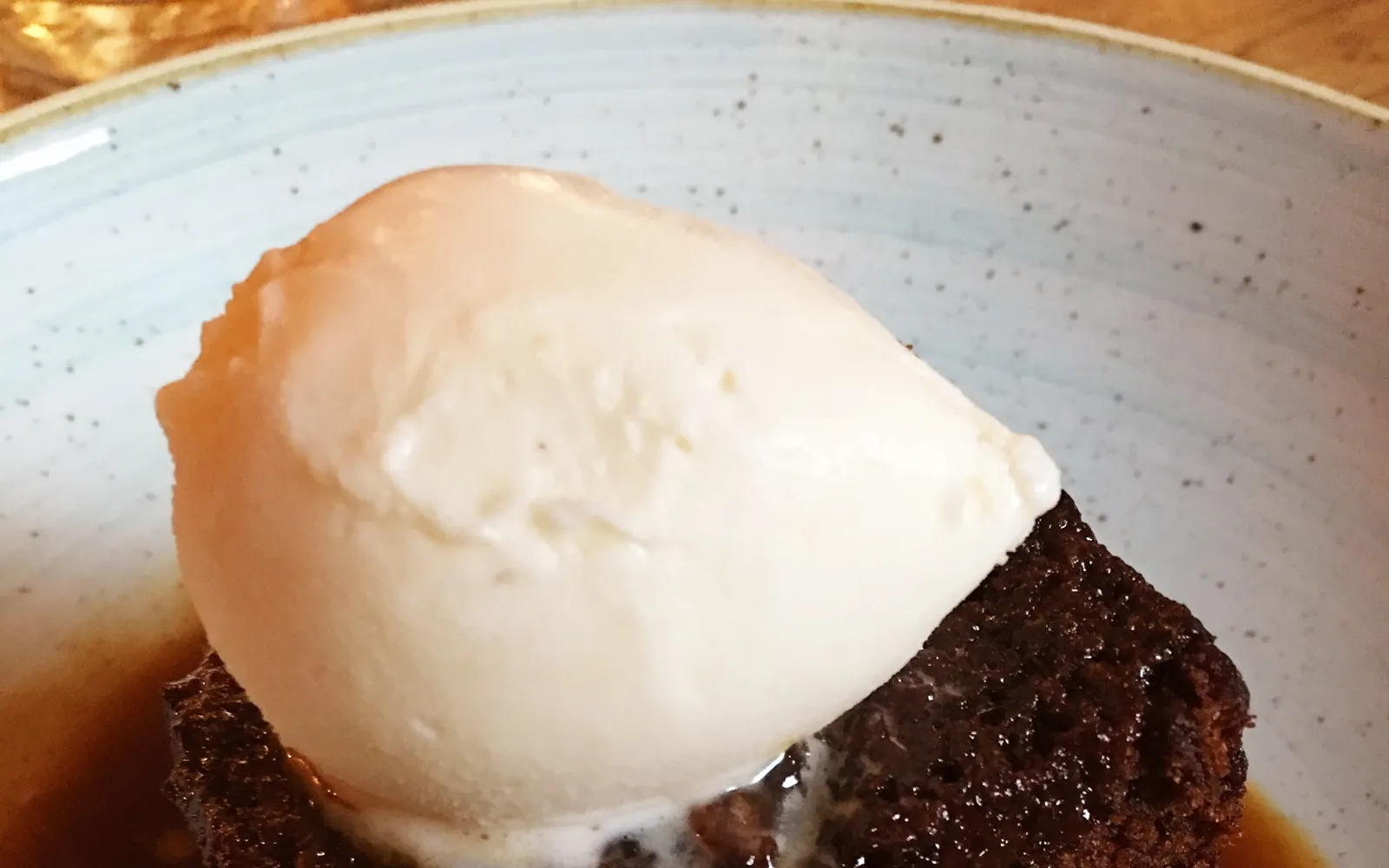 Sticky Toffee Pudding อบอุ่นสีน้ำตาลเข้มในจานขาว ซอส toffee สีน้ำตาลทองราดเต็มพร้อม ice cream วานิลลาขาวครีมวางข้างๆ ควันอ่อนๆ ลอยขึ้น