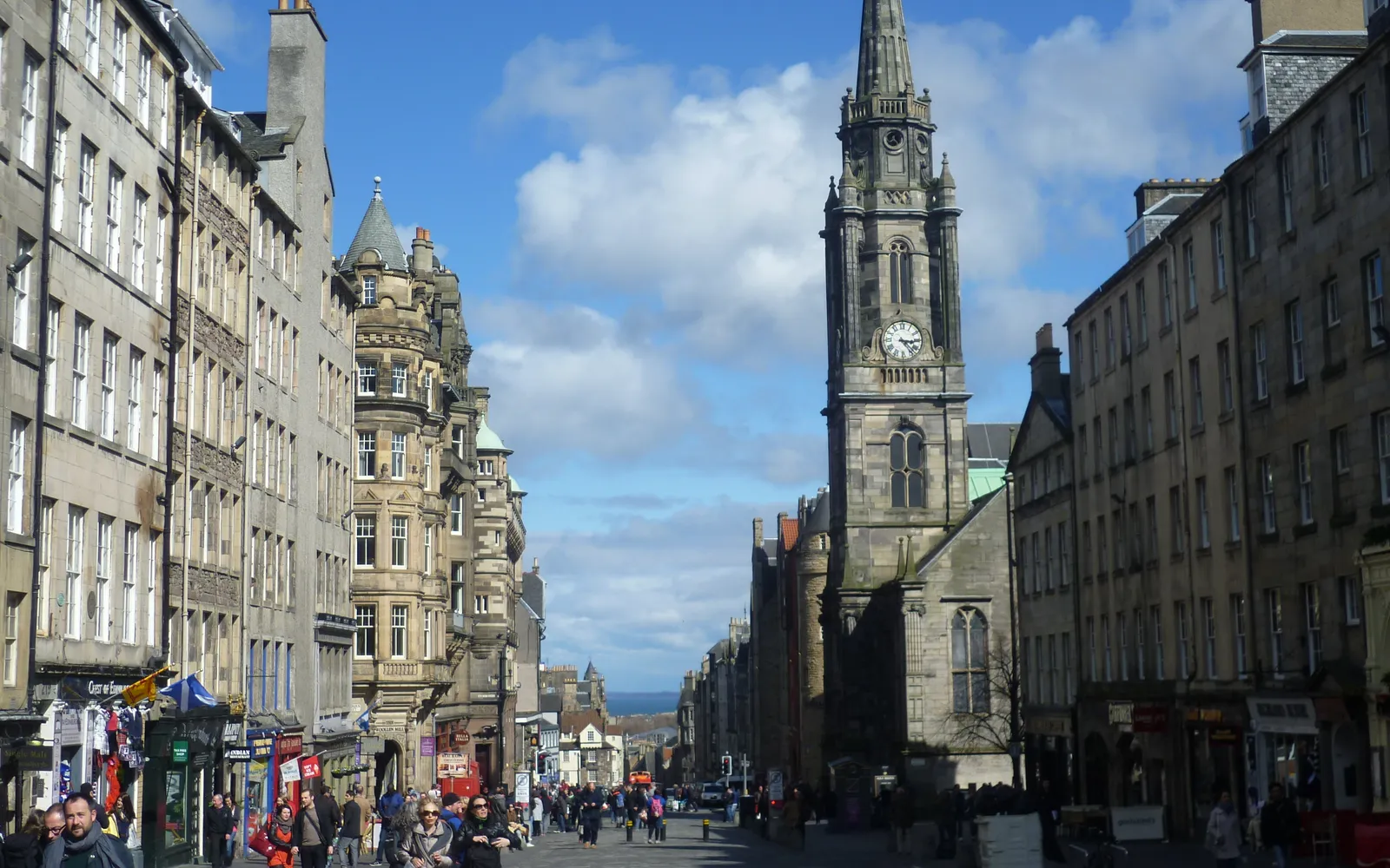 ถนน Royal Mile ในเอดินบะระ บ้านหินสีเทาเก่าแก่เรียงรายสองข้างทาง นักดนตรีเป่าแบกไปป์ในชุดสกอตแลนด์ยืนอยู่หน้าอาคารที่มีอายุหลายร้อยปี