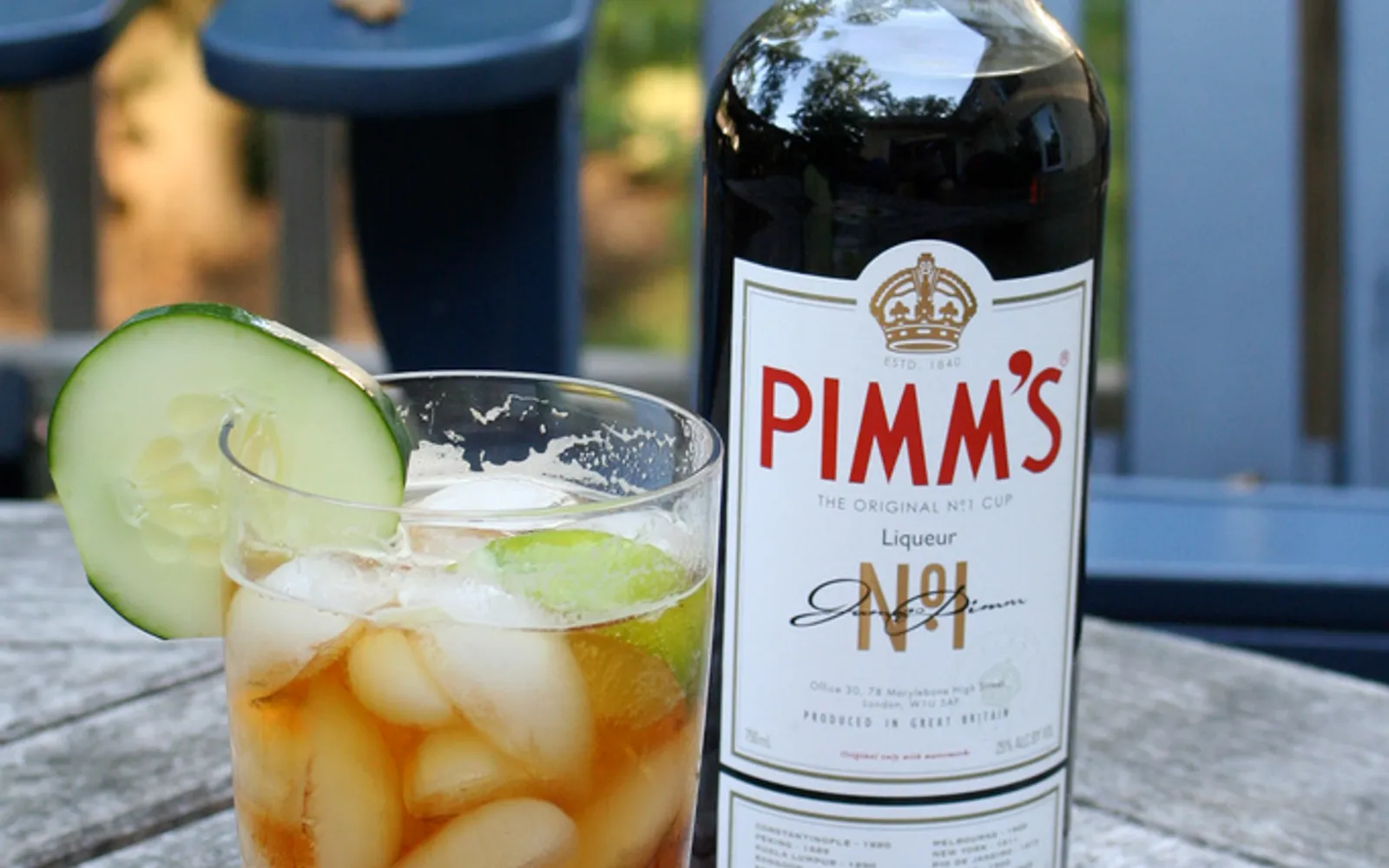 แก้ว Pimm Cup สูงใส น้ำดื่มสีน้ำตาลแดงใสแต่งด้วยแตงกวา สตรอว์เบอร์รี ใบมินต์ และน้ำแข็ง วางบนขอบผับริมแม่น้ำยามบ่าย
