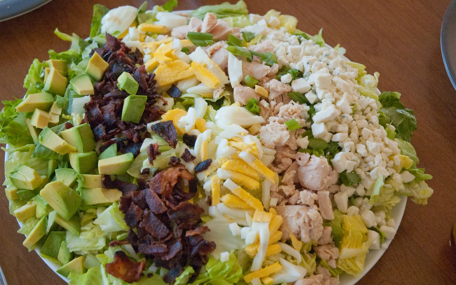 จาน Cobb Salad ขนาดใหญ่เรียงส่วนผสมเป็นแถว ไก่อก เบคอน ไข่ต้ม ชีส อาโวคาโด และมะเขือเทศบนผักกาดหอมสดเขียว